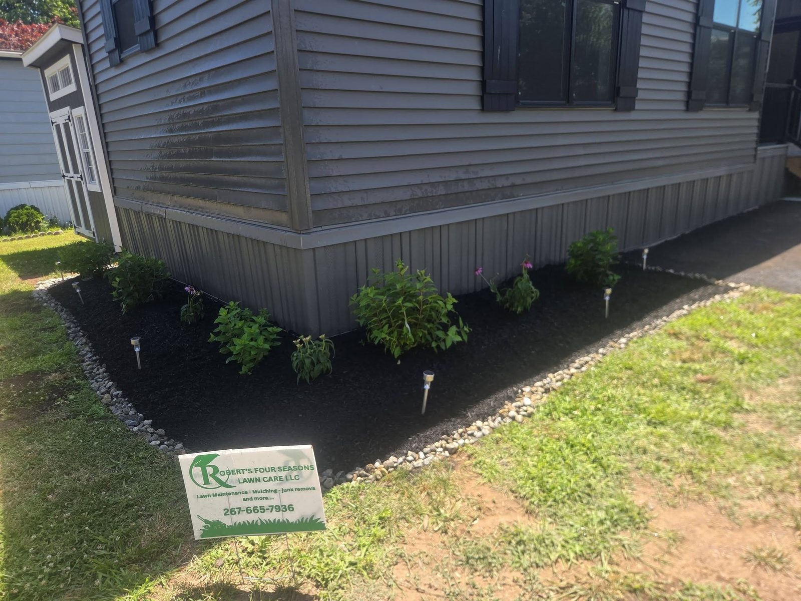 Foundation bed with black mulch, river rock border and solar path lights, with a Robert's Four Seasons yard sign